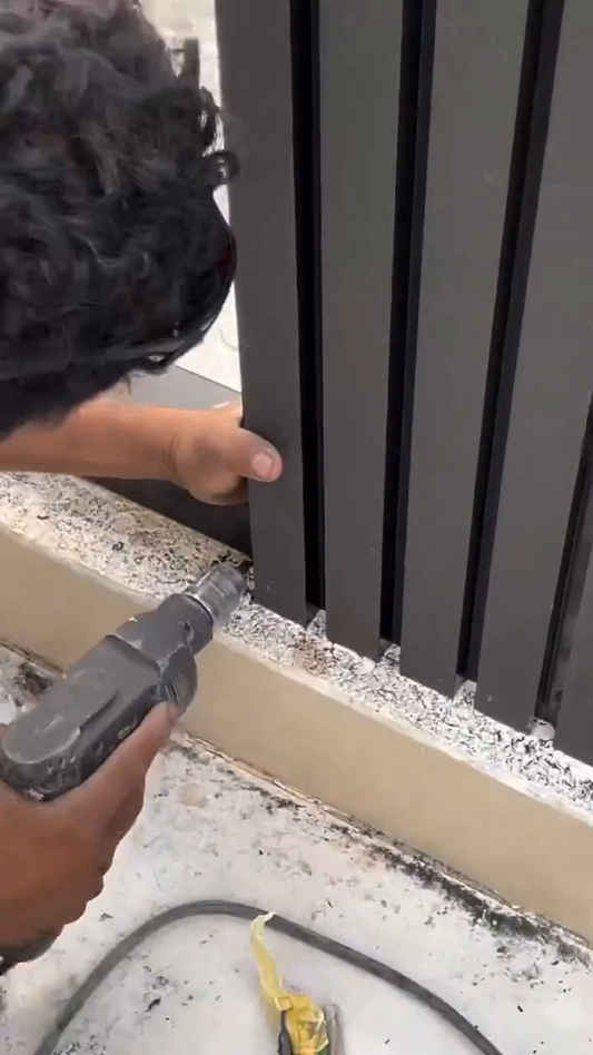 Instalador fijando listones verticales WPC de color oscuro sobre un muro exterior mediante perforación y anclaje.