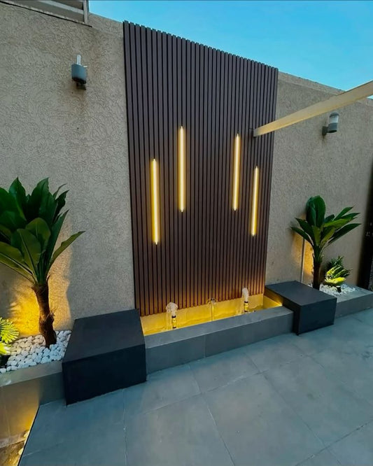 Muro exterior decorado con revestimiento WPC oscuro, luces LED verticales, fuente ornamental y jardineras con plantas.