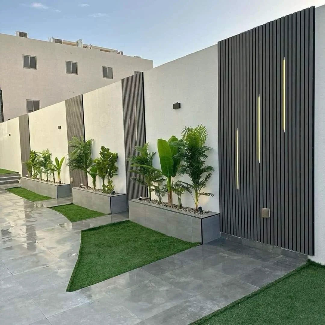 Muro exterior blanco decorado con paneles WPC negros, luces LED lineales y jardineras con plantas tropicales.