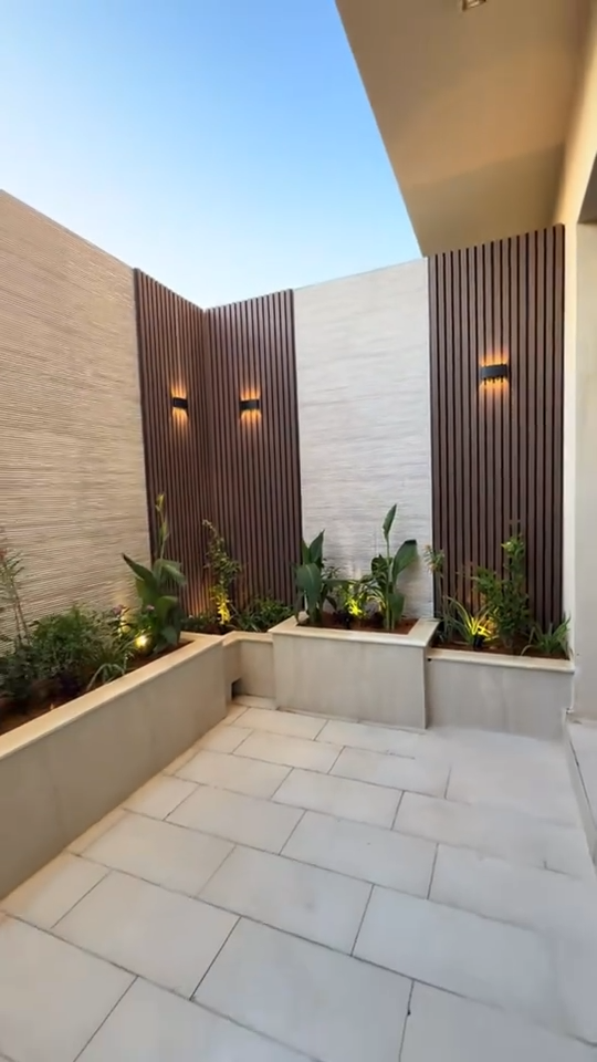 Patio moderno con paneles listonados WPC color madera, lámparas de pared, jardineras integradas y piso en tono claro.